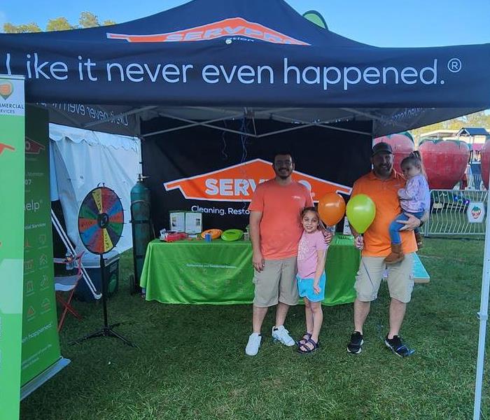 Two fathers stand under a black SERVPRO tent with their daughters holding SERVPRO balloons.
